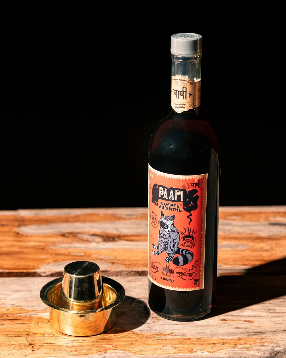 A bottle of Paapi Coffee Absinthe on a worn wooden table beside a brass coffee tumbler